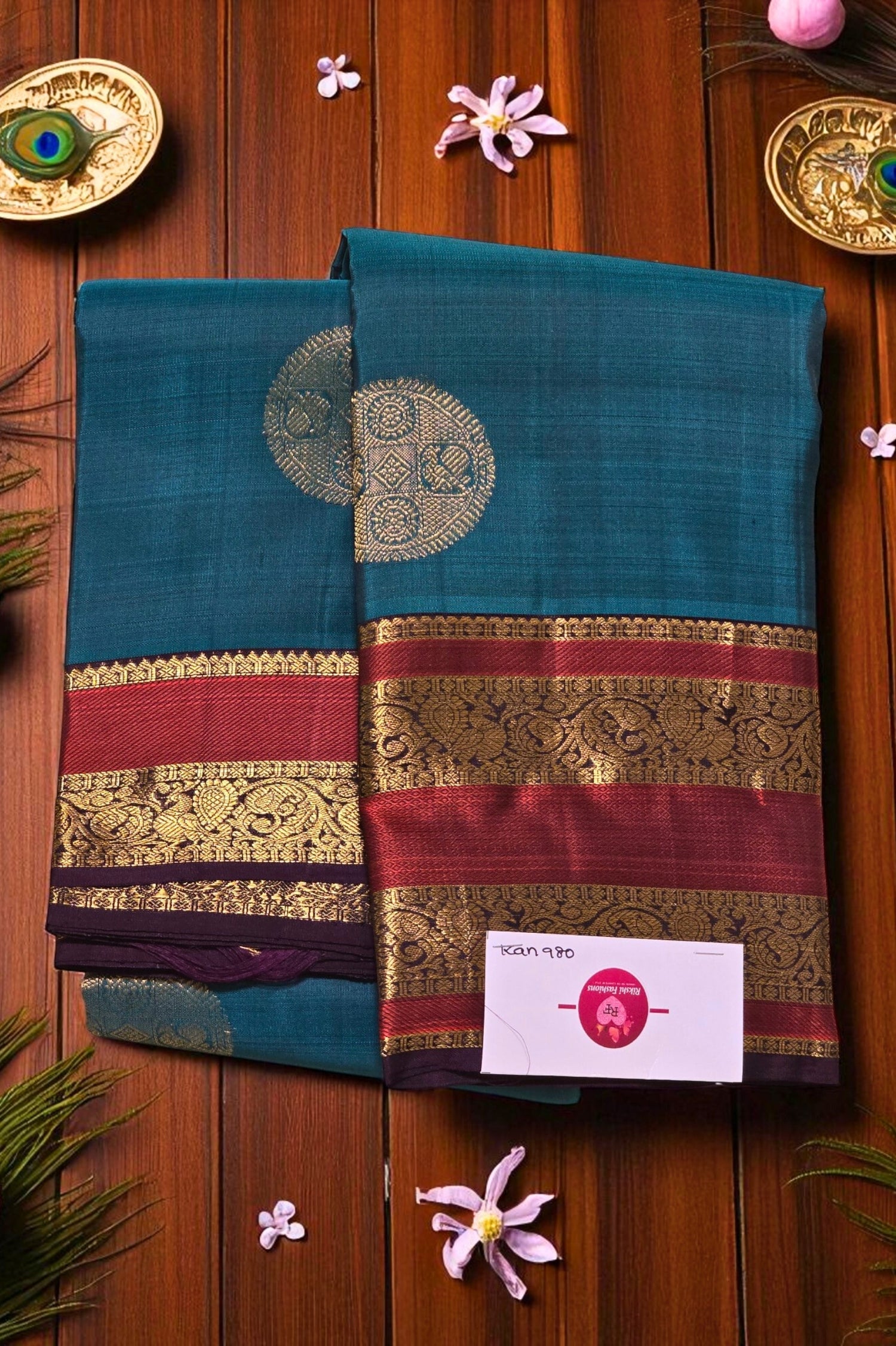 Two teal sarees with decorative borders on a wooden background with flowers and decorative items.