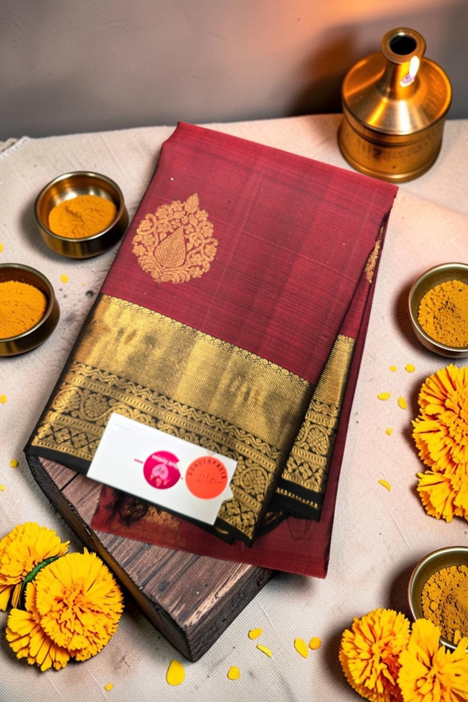 Maroon saree with gold border on a wooden board, surrounded by yellow flowers and small bowls.