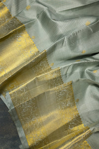 A close-up of a light sage green silk saree with a wide, ornate gold zari border and matching gold motifs woven throughout the body.