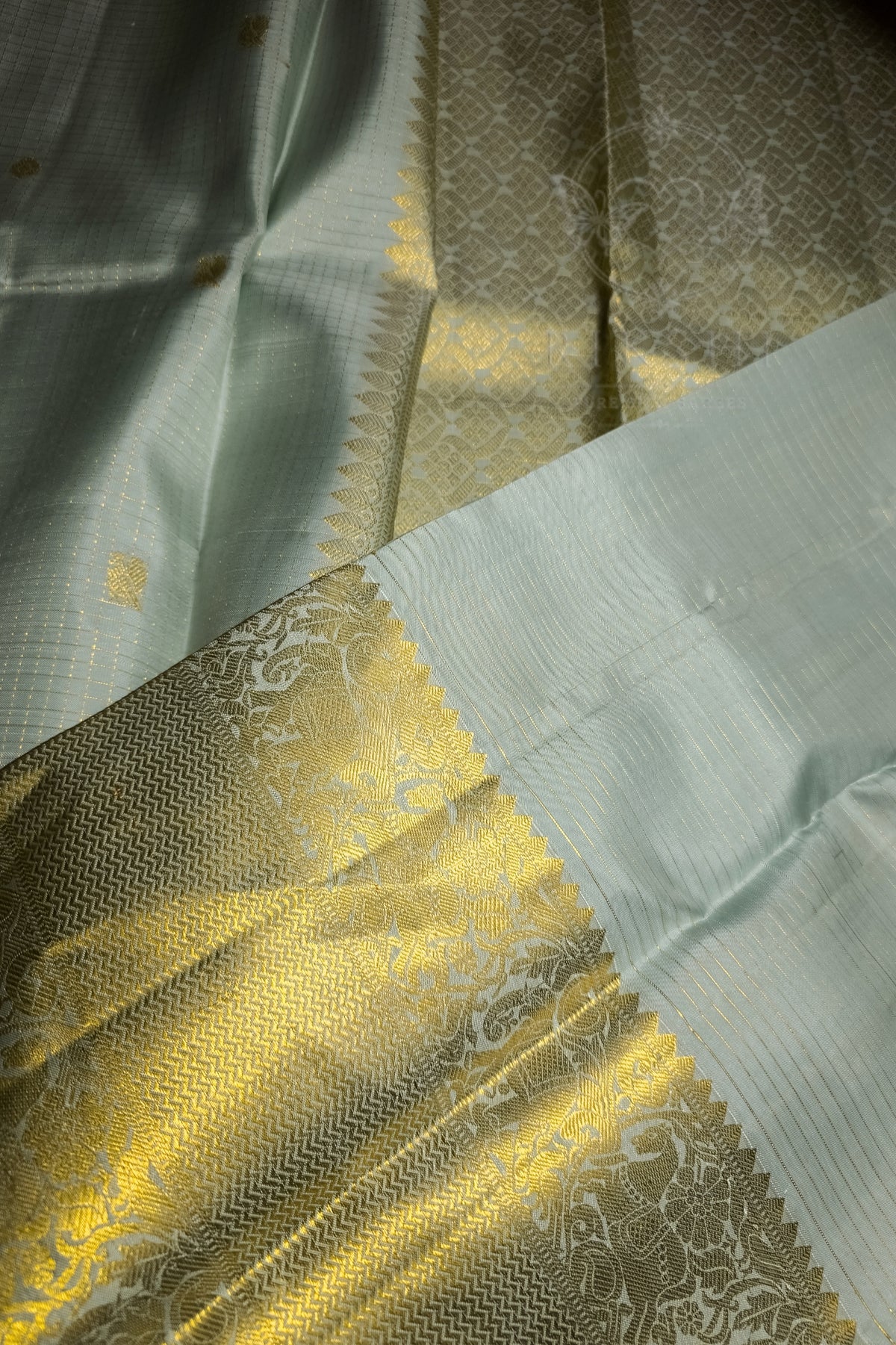 A close-up of a light sage green silk saree with a wide, ornate gold zari border and a white border with silver floral motifs.