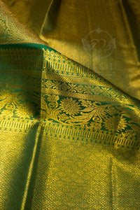 A close-up of a richly textured golden-yellow silk fabric, likely a sari, featuring intricate green and gold zari floral patterns and borders. A faint watermark logo for 'Rukshi' is visible in the center.