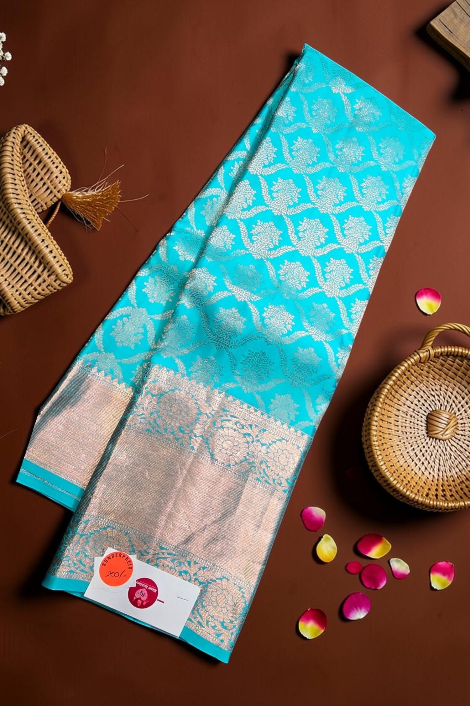 Turquoise and beige saree with floral patterns on a brown surface with wicker baskets and rose petals.