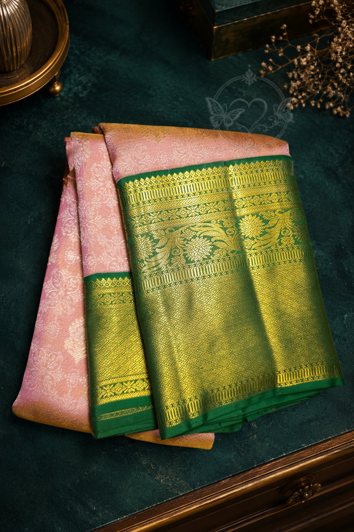 A folded pink and gold saree with a prominent green and gold zari border, displayed on a dark green textured surface.
