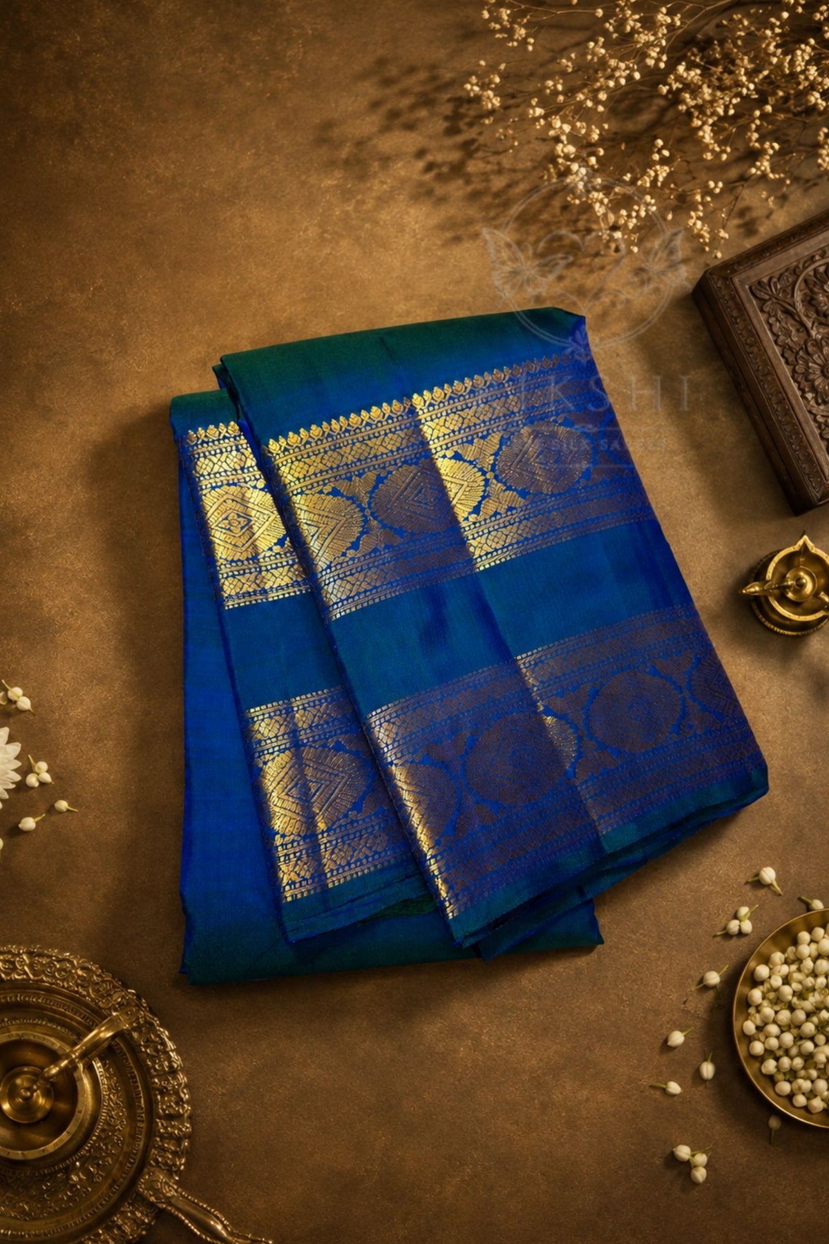 A folded royal blue and teal saree with intricate gold zari woven patterns, displayed on a textured brown background with traditional decorative props.
