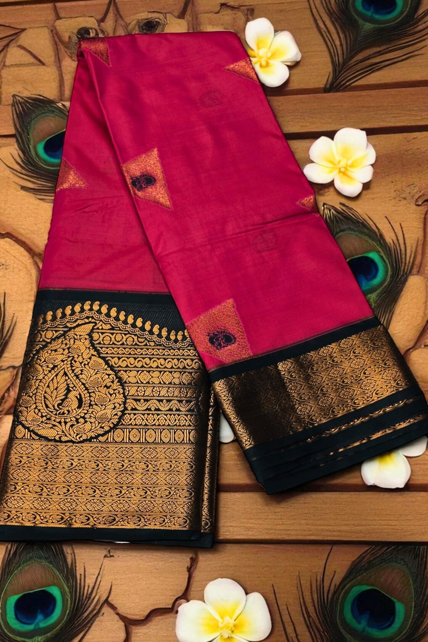 Pink saree with black and gold border on a wooden surface with peacock feathers and flowers.