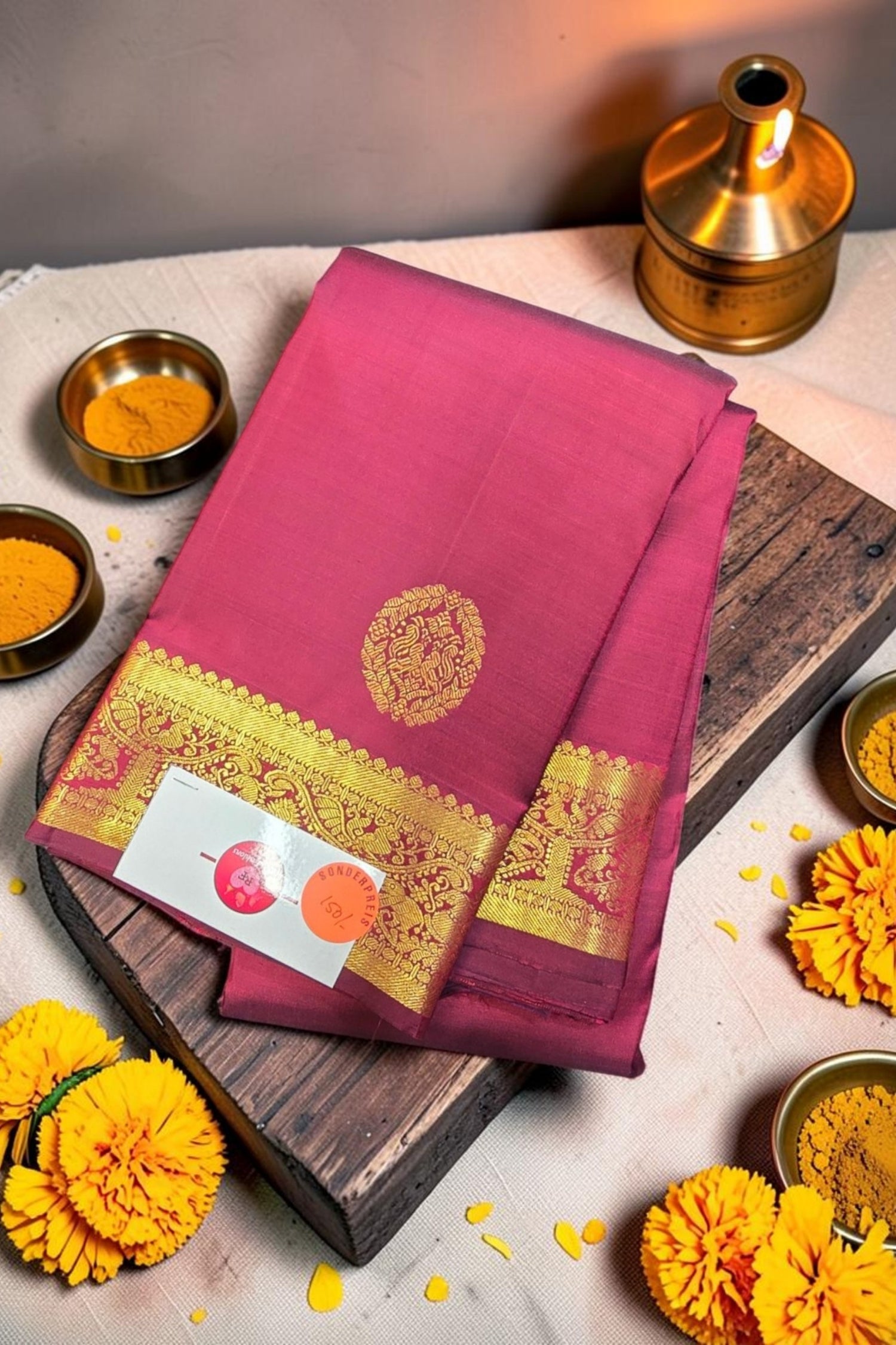 Pink saree with gold border on a wooden board, surrounded by yellow flowers and small containers.