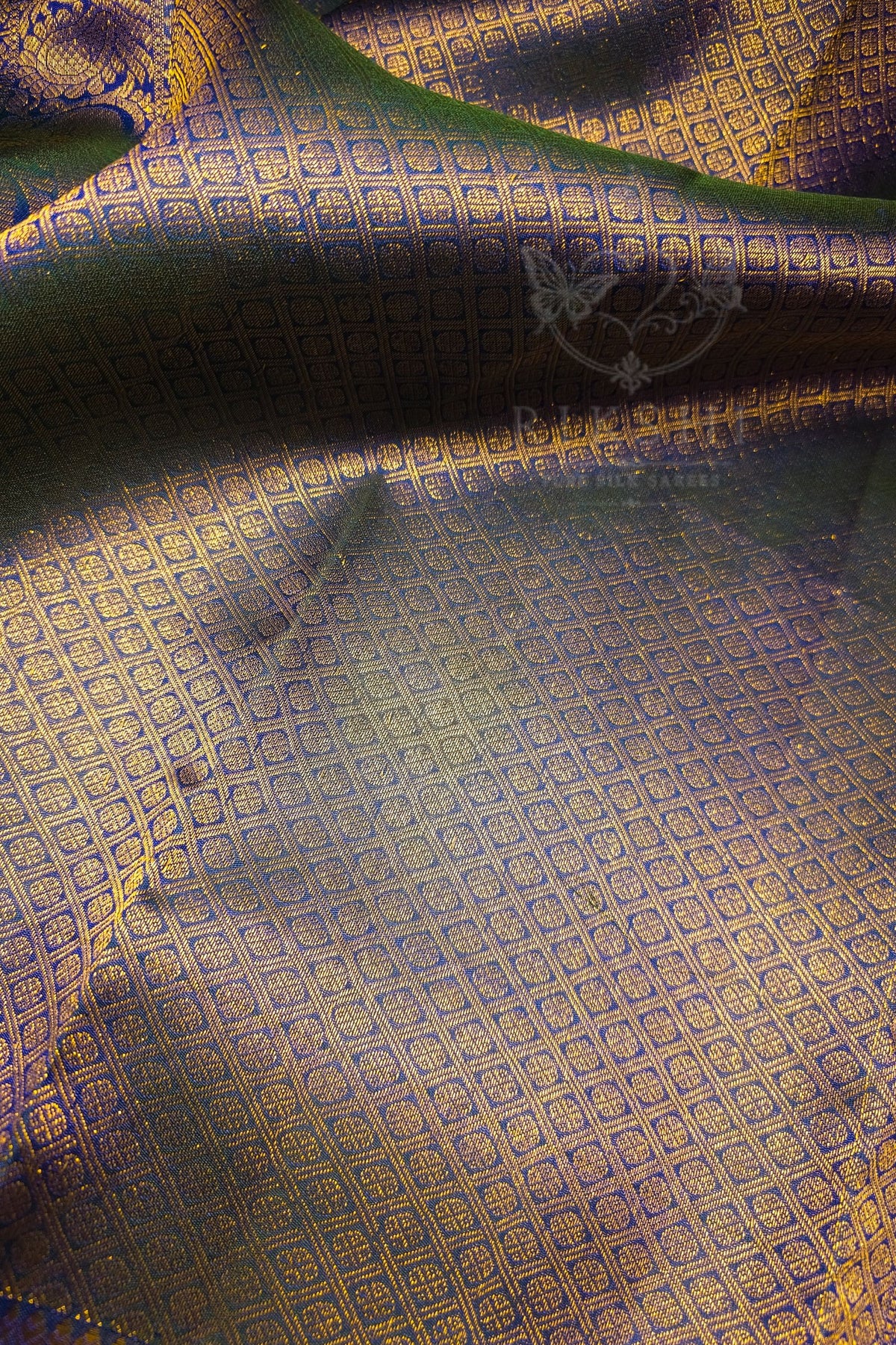 A close-up of a draped fabric with a repeating geometric woven pattern in gold and blue, showing folds and a subtle sheen.