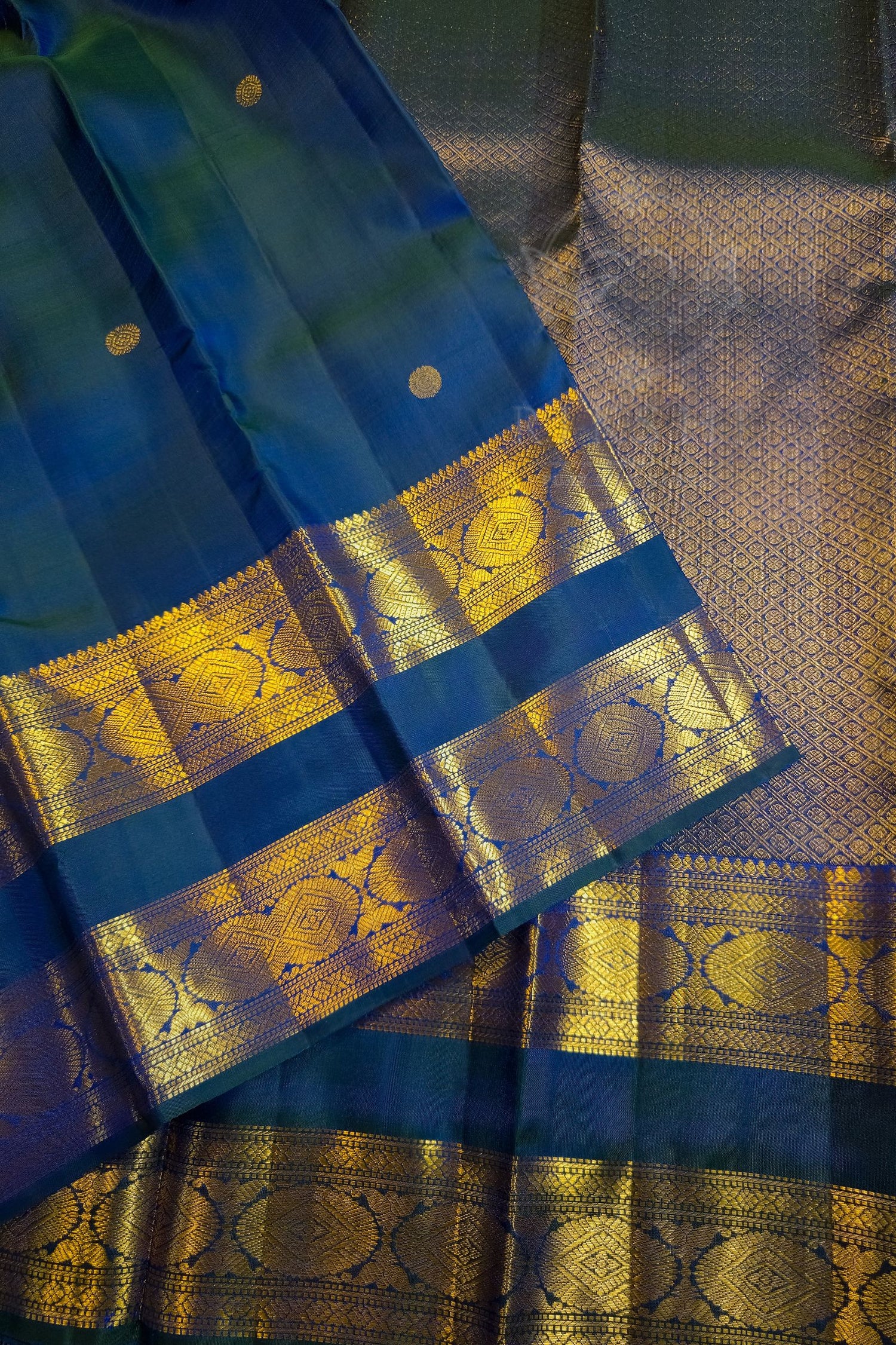 A close-up of a blue and gold woven fabric, likely a sari, featuring a rich metallic gold zari border with intricate circular motifs and a textured gold body.