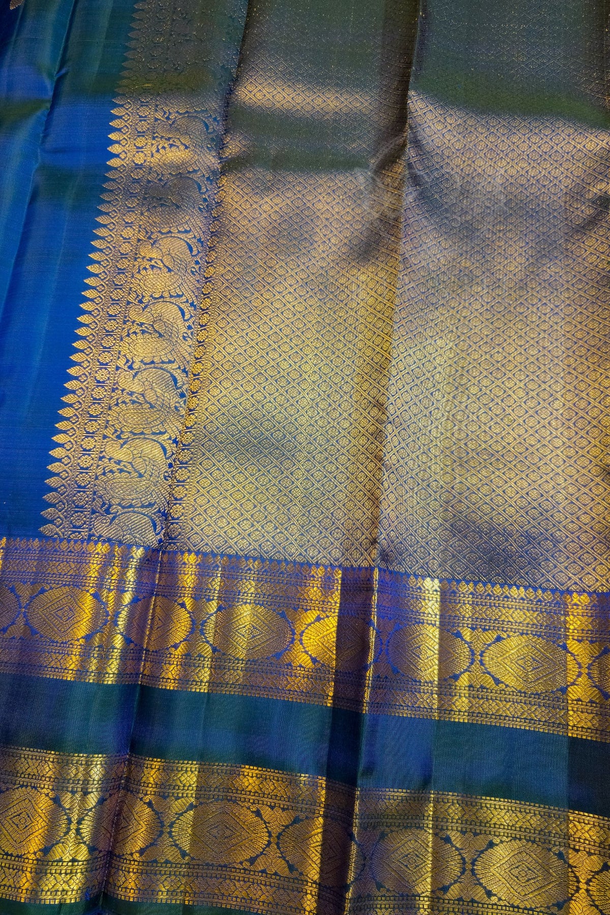 A close-up of a richly woven Indian silk saree, showcasing a metallic gold zari brocade pattern over a deep teal blue base.