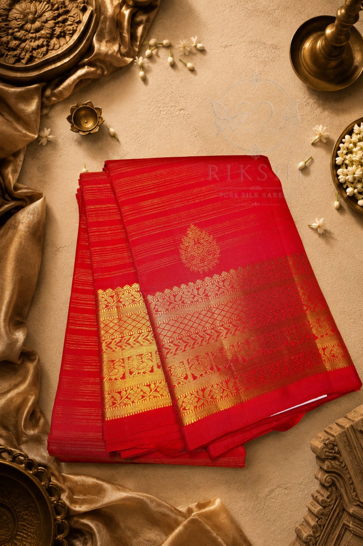 A folded red silk saree with intricate gold zari woven patterns and a floral motif, displayed on a textured beige surface with traditional decorative props.