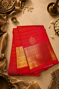 A folded red silk saree with intricate gold zari woven patterns and a floral motif, displayed on a textured beige surface with traditional decorative props.