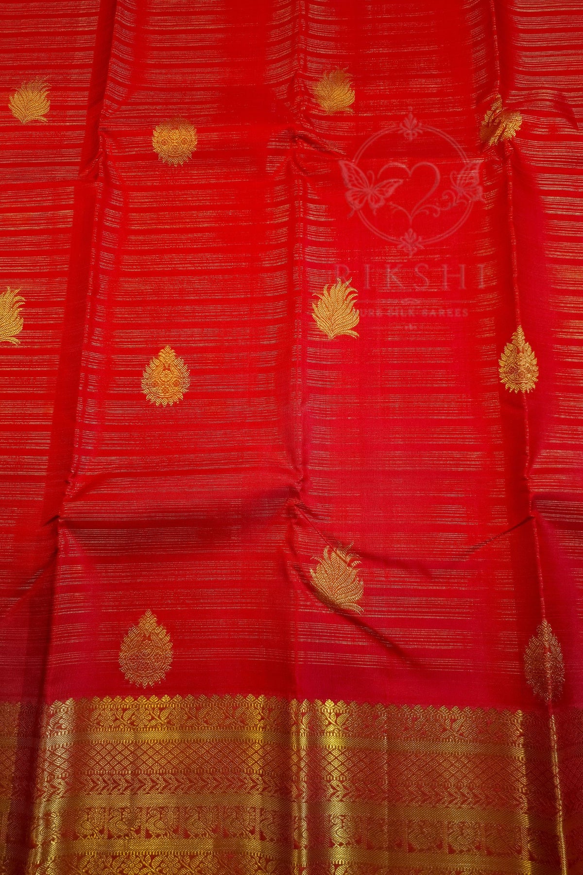A close-up of a red silk saree with gold zari embroidery, featuring small teardrop motifs and a wide decorative gold border at the bottom. A faint watermark logo for 'Kshiti' is visible in the center.
