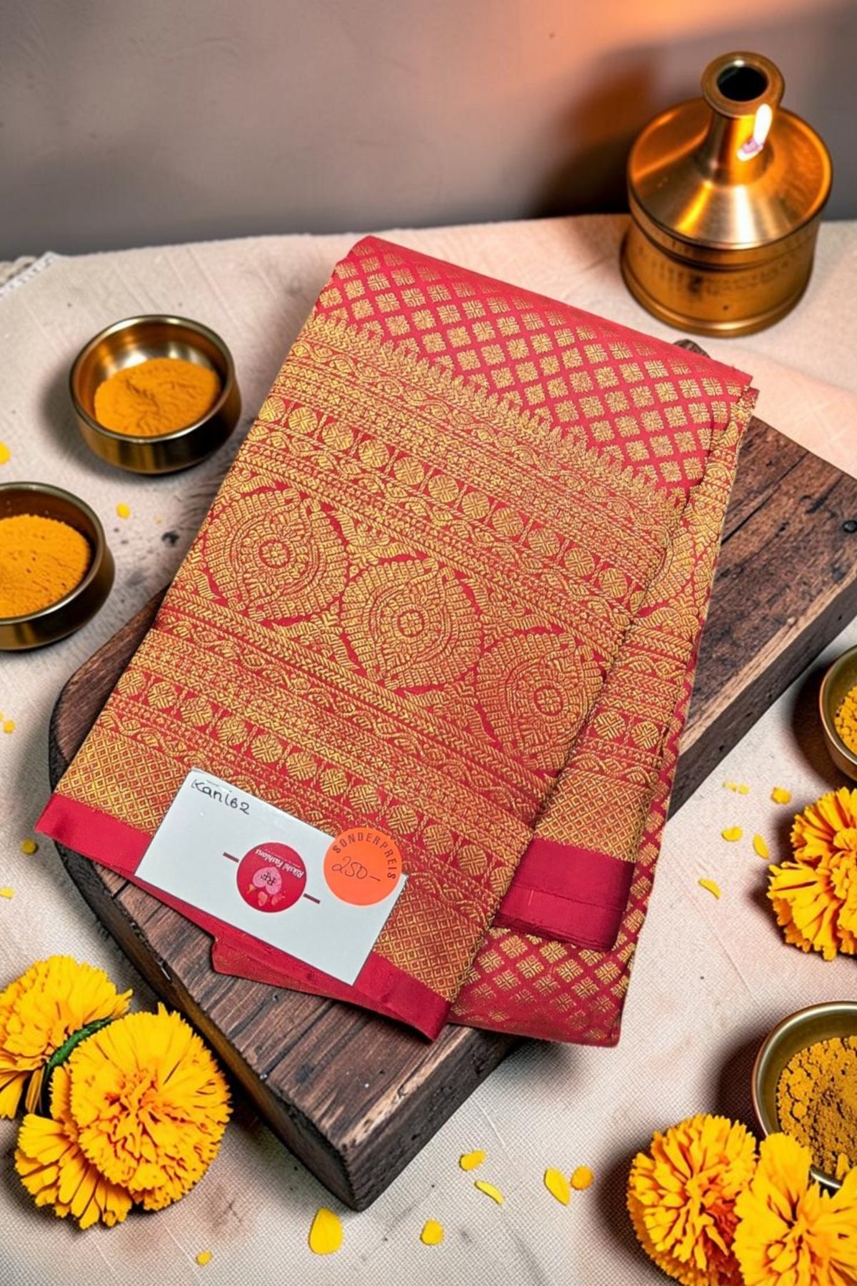 Red and gold patterned fabric with a label on a wooden board, surrounded by yellow flowers and small bowls.