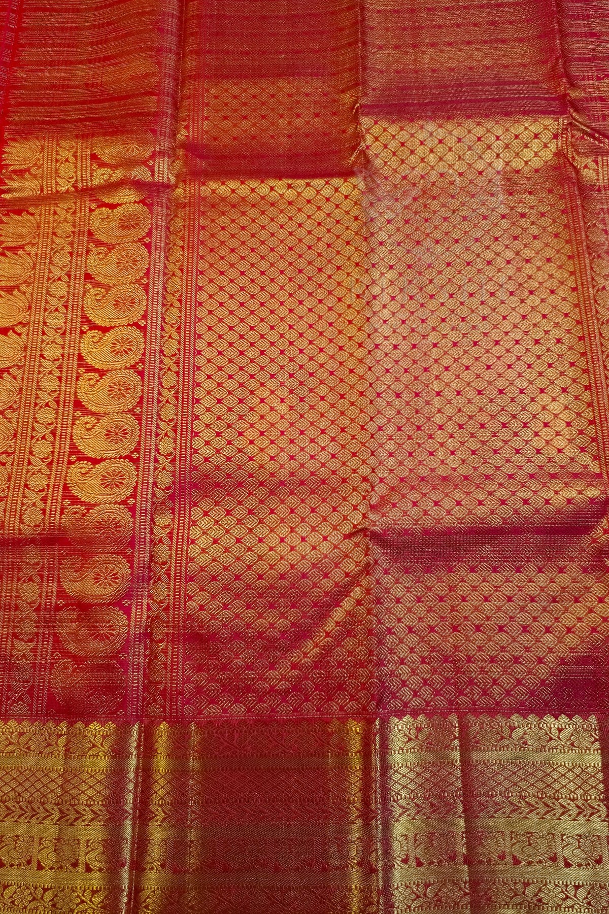 A close-up of a red and gold woven fabric, likely a silk saree or textile, featuring intricate metallic zari patterns and borders.