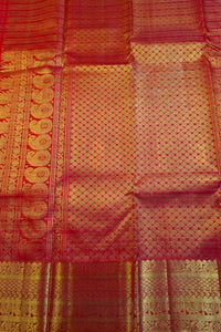 A close-up of a red and gold woven fabric, likely a silk saree or textile, featuring intricate metallic zari patterns and borders.
