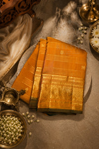 An orange-gold silk saree with intricate gold zari embroidery, folded neatly and styled on a textured gray surface with traditional brass props and white flowers.