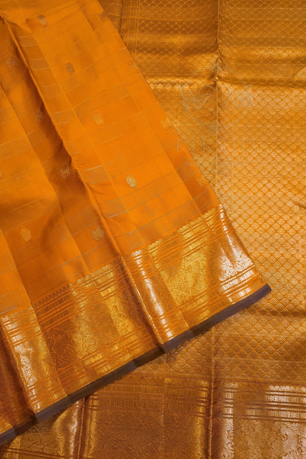 A close-up of an orange-gold silk saree with metallic gold zari embroidery, showing the woven pattern and border details.