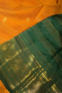 A close-up of a richly patterned saree, showing a vibrant orange body with subtle gold motifs and a wide dark green border adorned with intricate gold zari weaving.