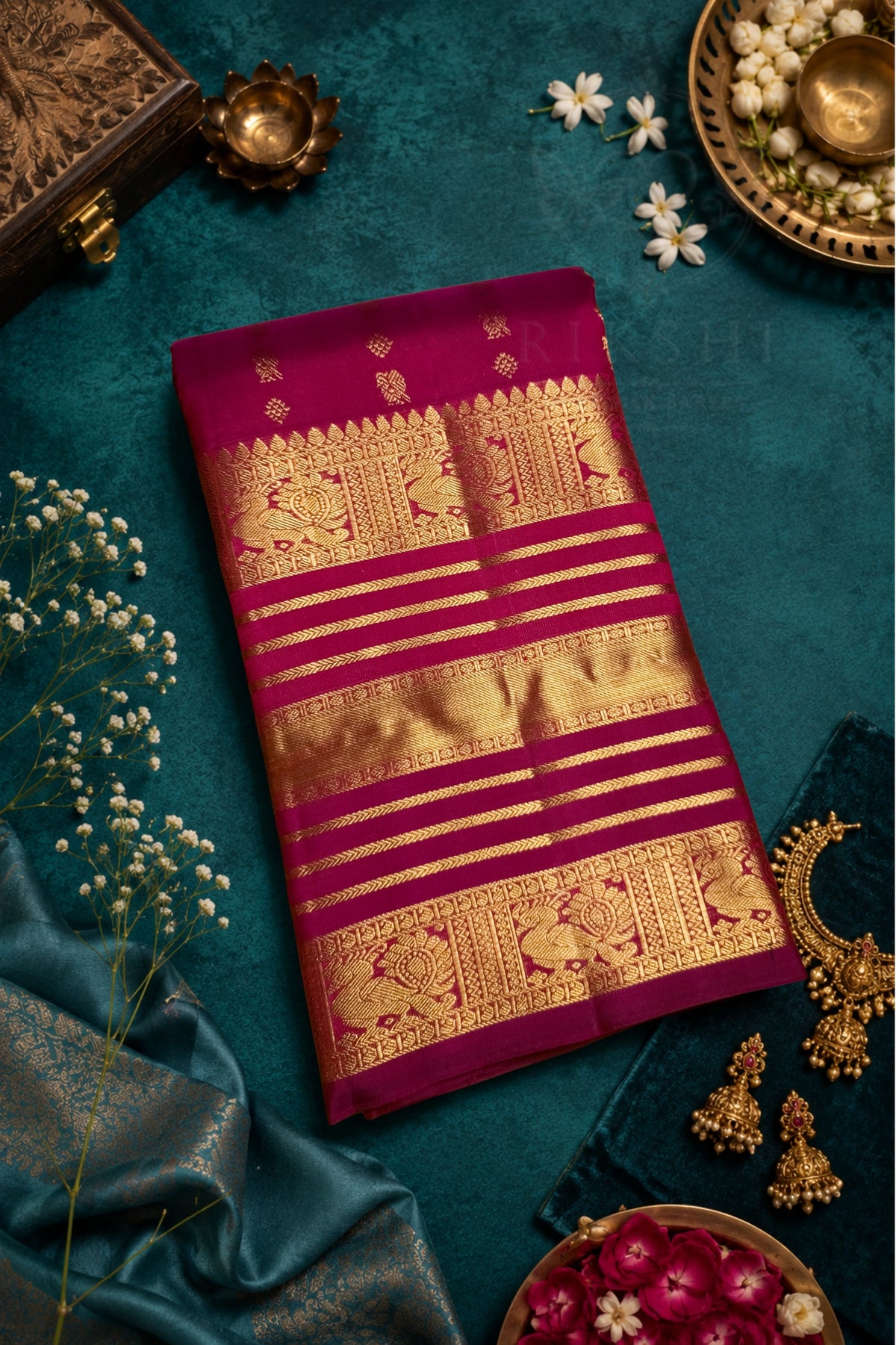 A folded deep magenta silk saree with intricate gold zari brocade patterns, laid flat on a dark teal textured background surrounded by decorative props including white flowers, a brass pot, and a blue satin fabric.