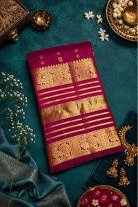 A folded deep magenta silk saree with intricate gold zari brocade patterns, laid flat on a dark teal textured background surrounded by decorative props including white flowers, a brass pot, and a blue satin fabric.