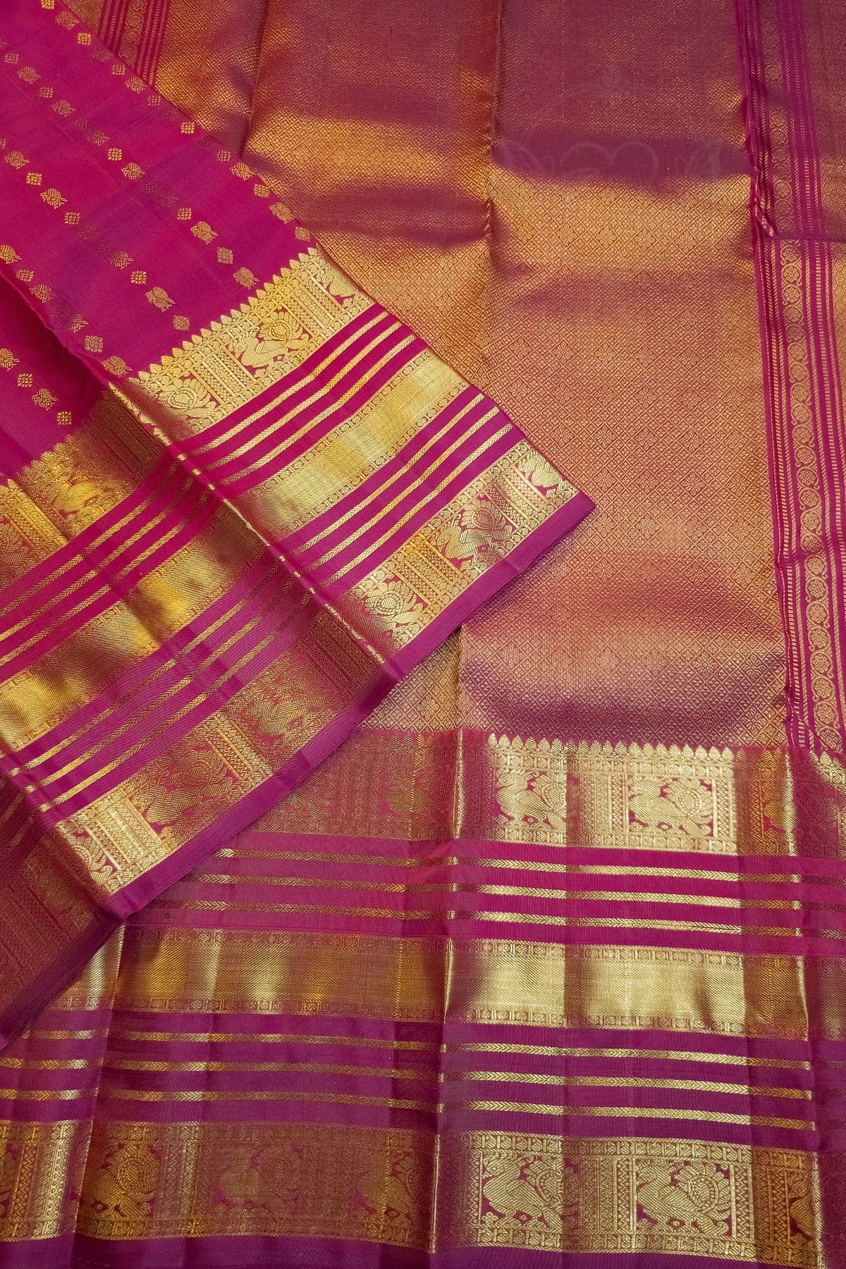 A close-up of a richly patterned silk saree, showcasing its golden zari brocade work, pink and gold stripes, and a vibrant magenta border.