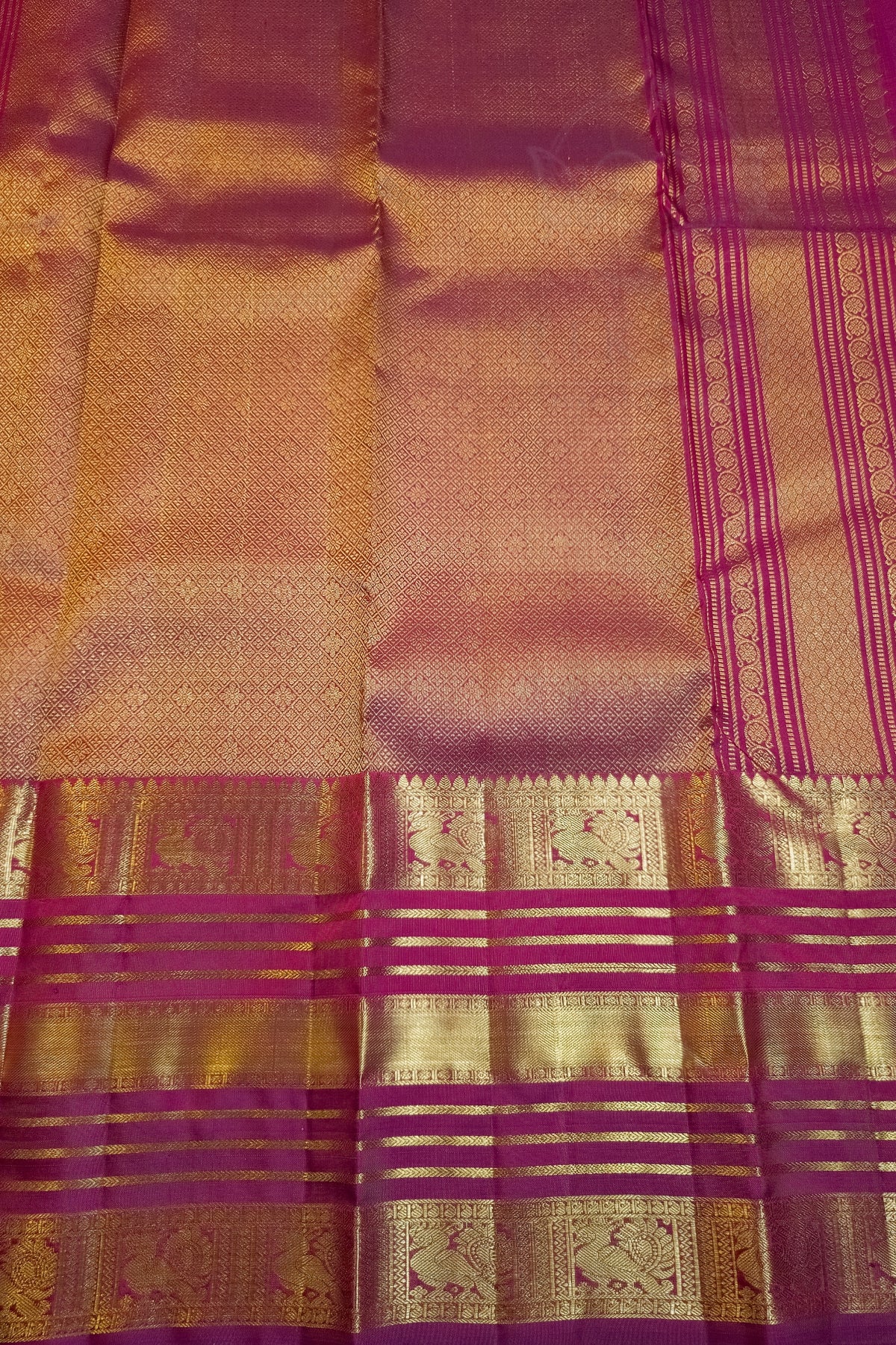 A close-up of a woven textile, likely a sari, featuring a warm pinkish-gold body with metallic gold zari patterns and magenta-purple borders.