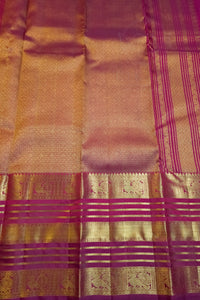 A close-up of a woven textile, likely a sari, featuring a warm pinkish-gold body with metallic gold zari patterns and magenta-purple borders.