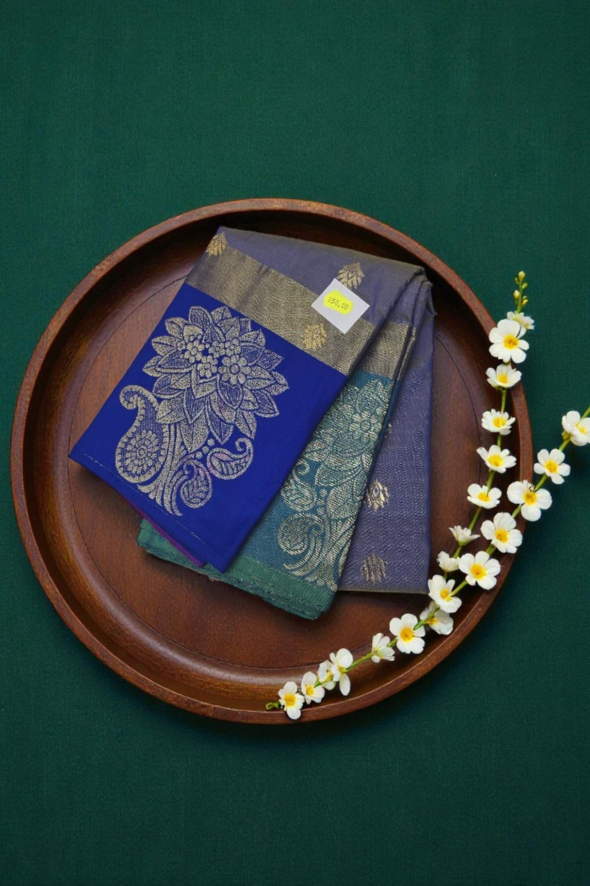 Three embroidered cloths on a wooden tray with a green background and white flowers.