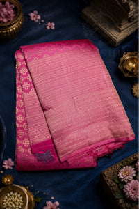 A folded pink Banarasi silk saree with intricate gold zari brocade patterns, displayed on a dark blue fabric background with decorative brass and floral props.