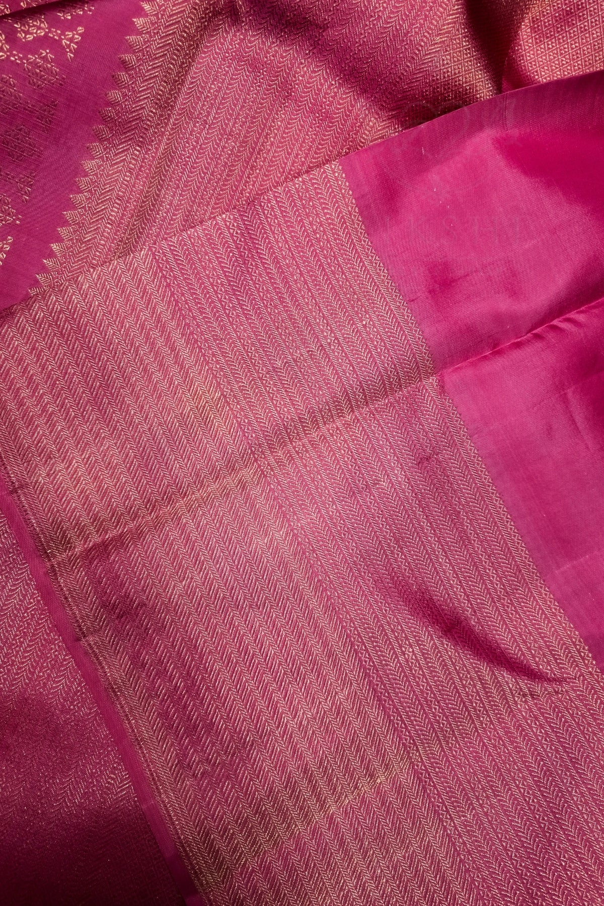 A close-up of a pink silk saree or fabric, showing the woven body with fine gold zari detailing and a solid pink border.