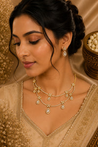 Woman wearing a gold necklace and earrings with a beige outfit against a warm-toned background.