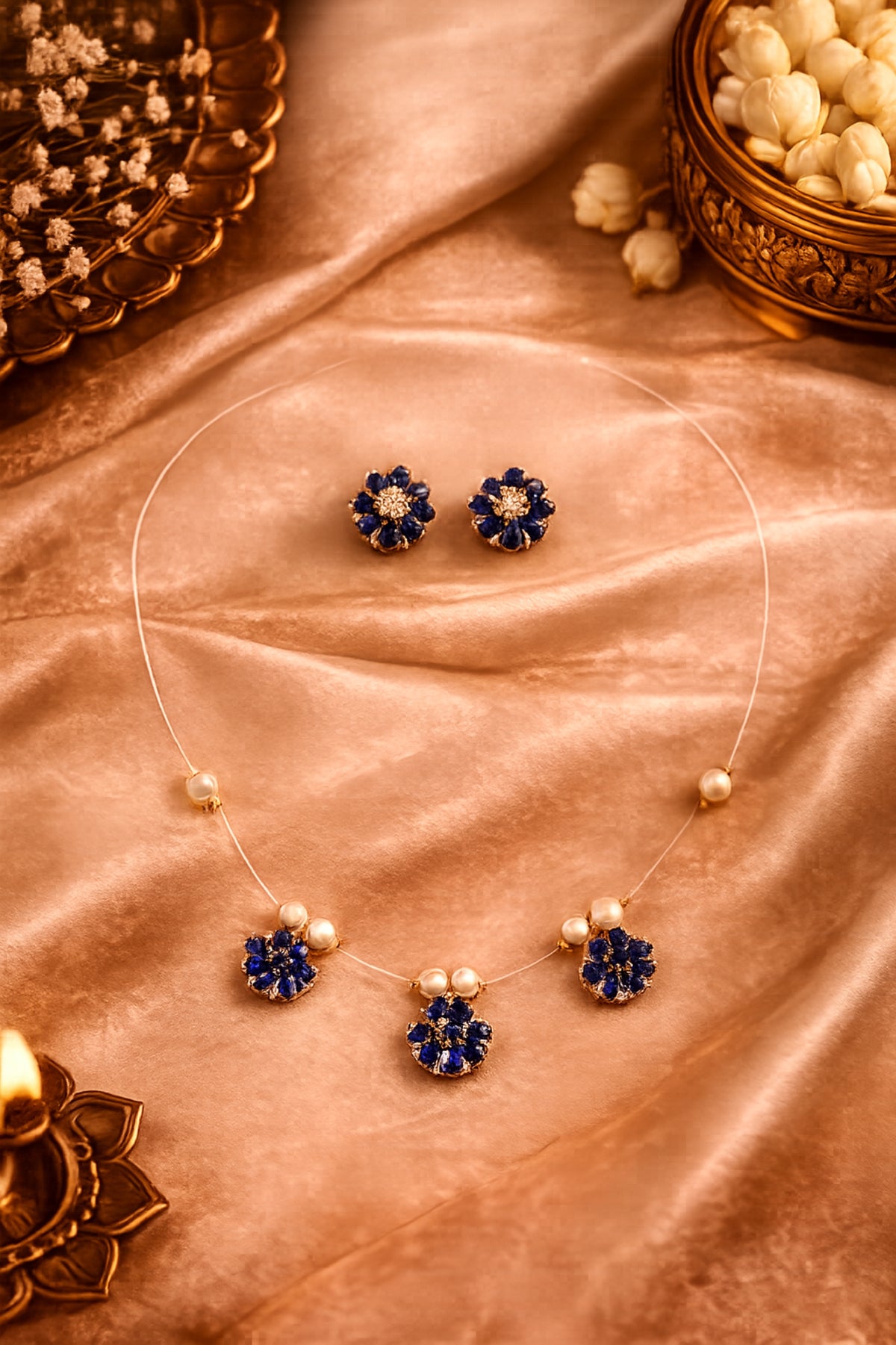 A gold jewelry set featuring a necklace and matching earrings with deep blue flower-shaped stones and small pearl-like accents, arranged on draped peach satin fabric with decorative props.