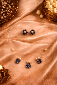 A gold jewelry set featuring a necklace and matching earrings with deep blue flower-shaped stones and small pearl-like accents, arranged on draped peach satin fabric with decorative props.