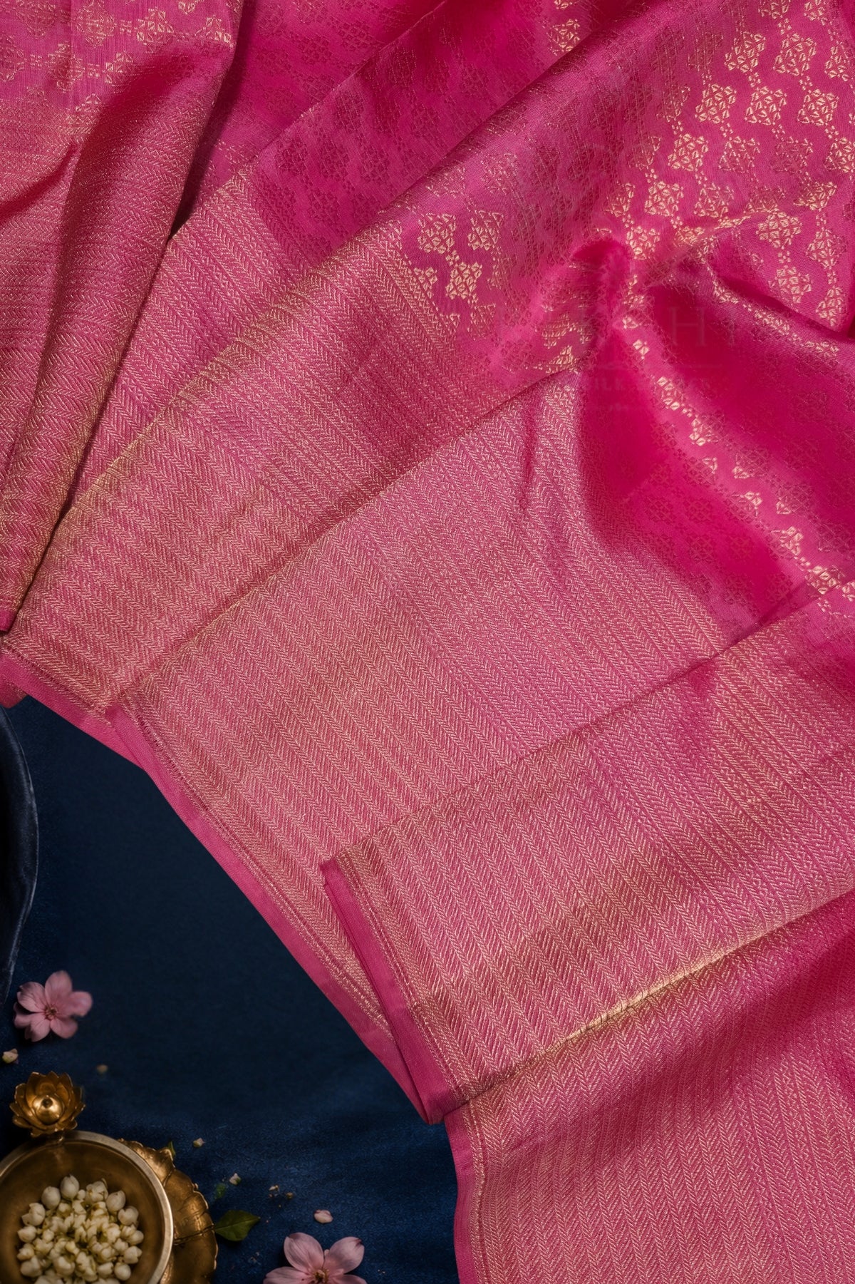 A close-up of a pink silk saree with gold zari woven patterns and borders, draped elegantly over a dark blue fabric surface with small decorative flowers and a brass bowl in the corner.