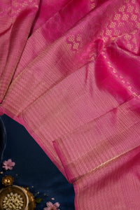 A close-up of a pink silk saree with gold zari woven patterns and borders, draped elegantly over a dark blue fabric surface with small decorative flowers and a brass bowl in the corner.
