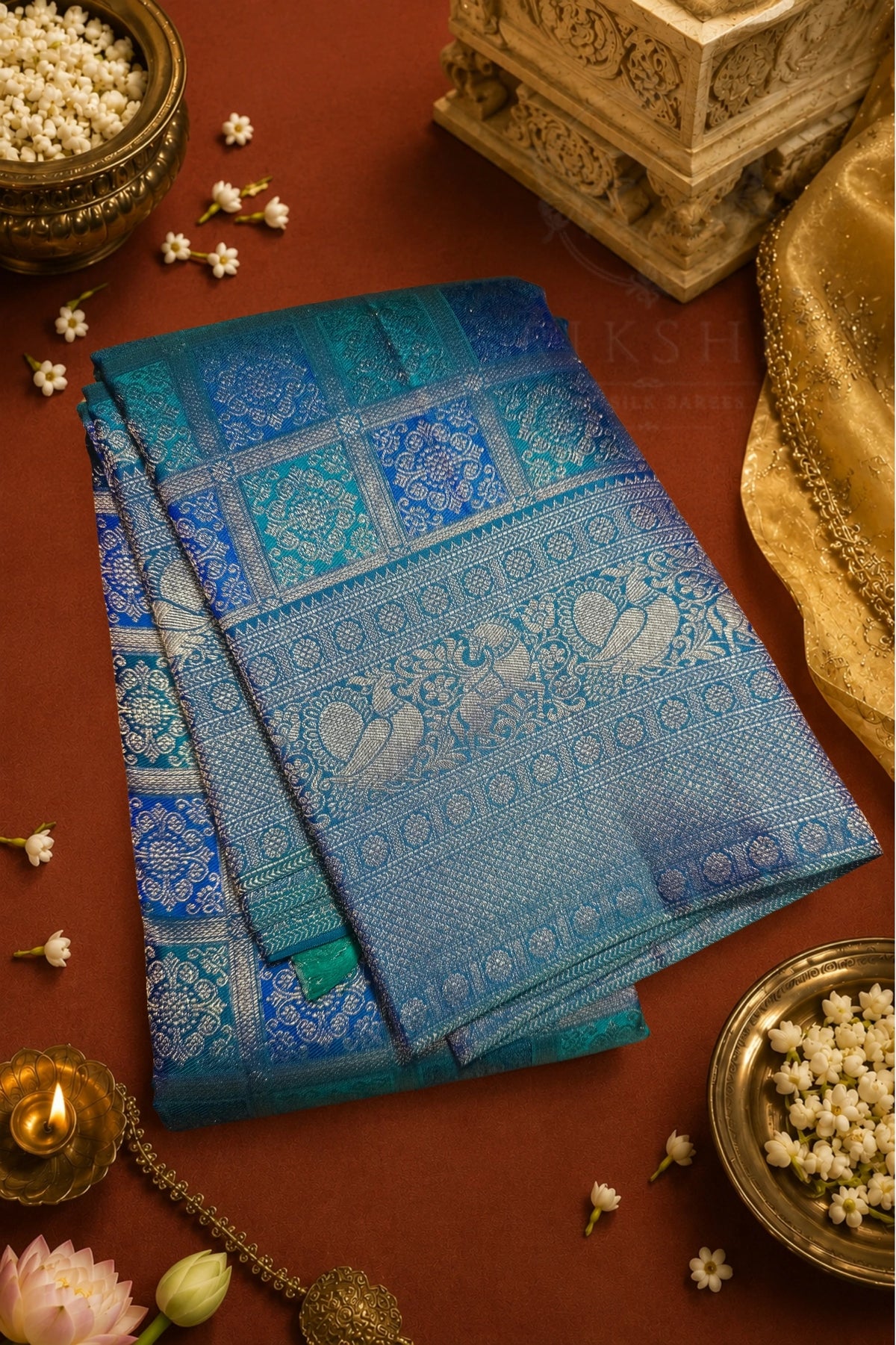 A folded blue and teal Banarasi silk saree with intricate silver zari embroidery, displayed on a warm brown background with traditional decorative props.