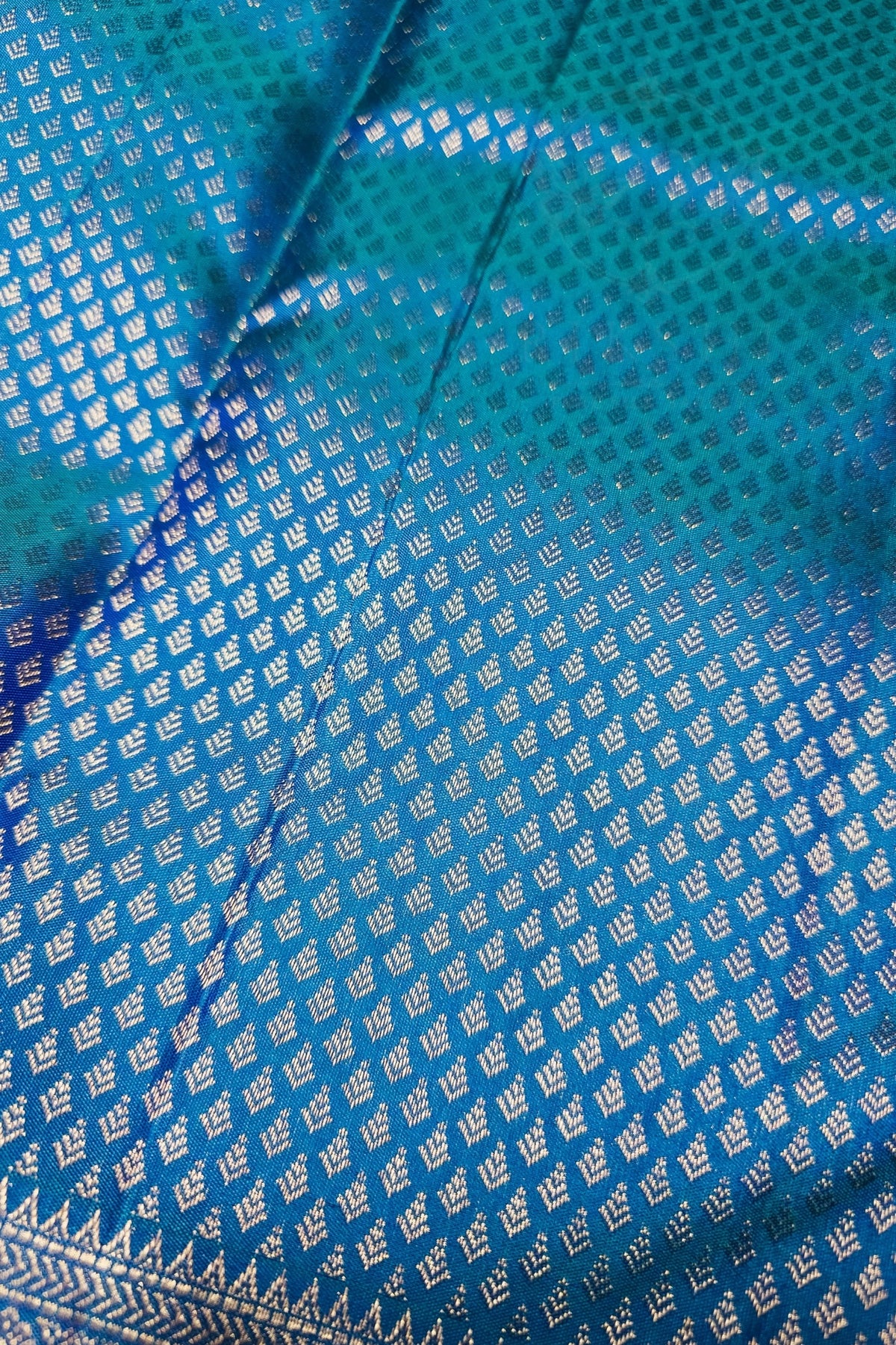 A close-up of a blue and teal patterned fabric with metallic silver woven motifs and a decorative border.
