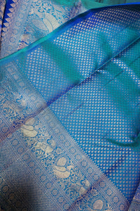 A close-up of a blue and teal silk saree fabric with intricate silver zari embroidery, featuring a detailed border and a checkered pattern.