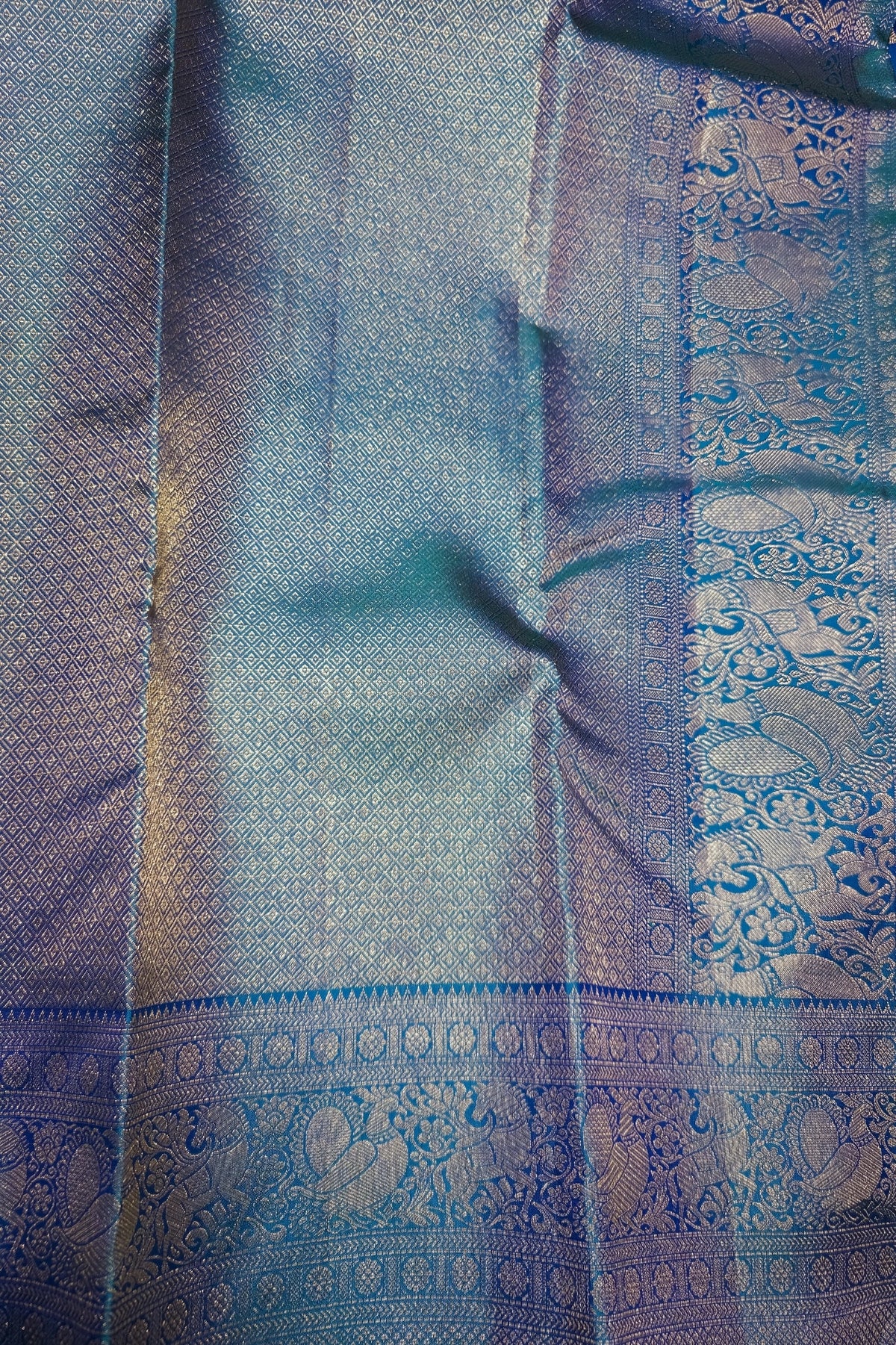 A close-up of a woven fabric, likely a silk saree or textile, featuring a shimmering blue and silver body with an ornate metallic border in blue and gold tones.