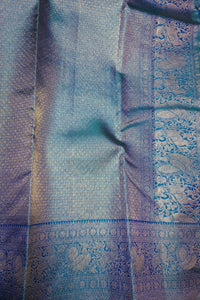 A close-up of a woven fabric, likely a silk saree or textile, featuring a shimmering blue and silver body with an ornate metallic border in blue and gold tones.