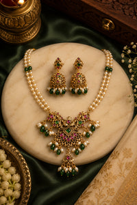 A traditional Indian jewelry set featuring a pearl necklace and matching earrings with gold-plated jhumka-style pendants adorned with green and pink stones, displayed on a round marble stand.