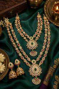 An ornate gold-toned traditional Indian jewelry set displayed on rich emerald green silk fabric, featuring a layered necklace, matching earrings, and decorative props including a lit diya and wooden box.