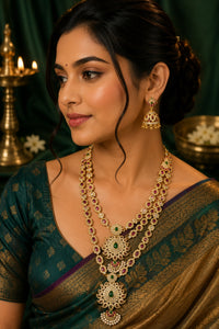 A woman wearing an ornate layered gold necklace with green and red stones, matching dangling earrings, and a green sari with gold zari detailing, posed against a dark green draped background with warm decorative lighting.
