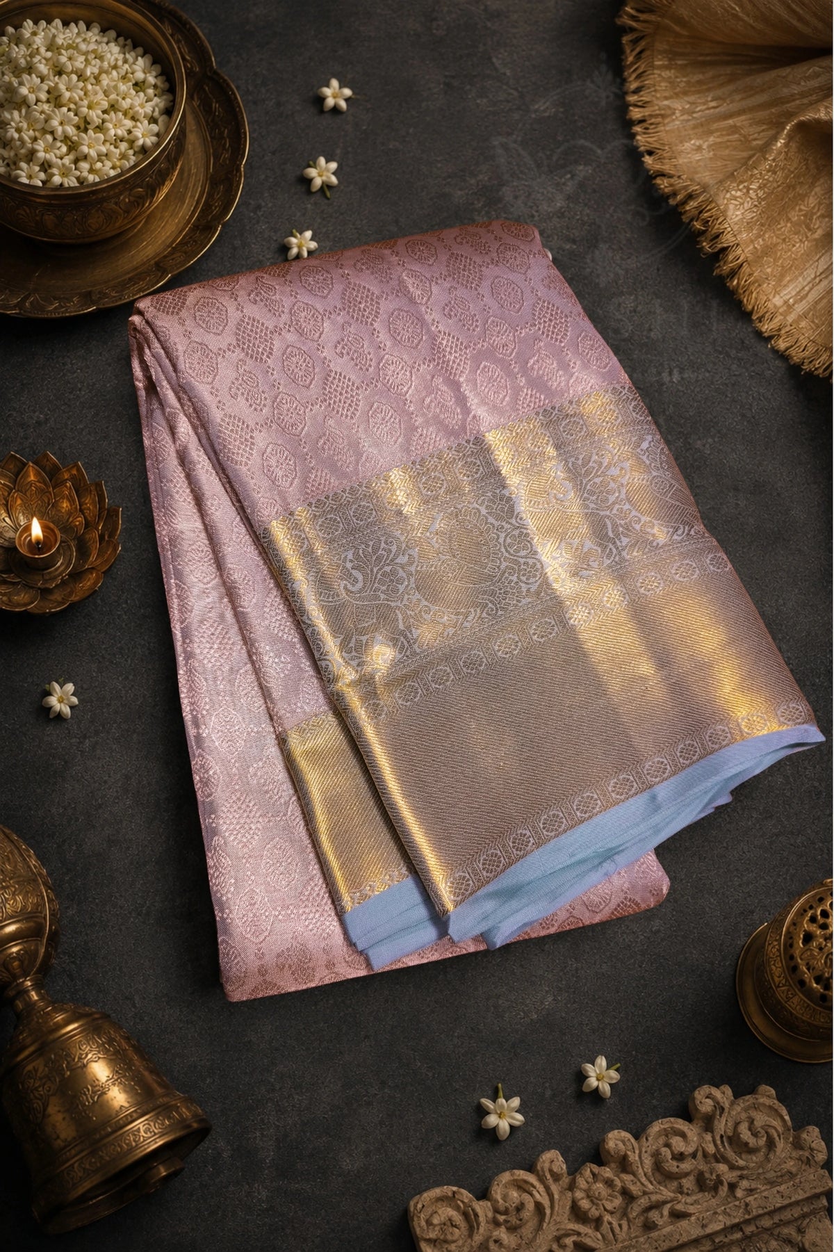 A folded pink and gold brocade saree with a light blue inner layer, styled on a dark textured background with traditional Indian decorative props.