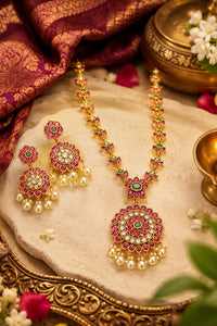 Gold-toned Indian jewelry set featuring a necklace and matching earrings with pink and green stone floral motifs, pearl bead drops, and a central green stone, displayed on a stone slab with traditional decorative props.