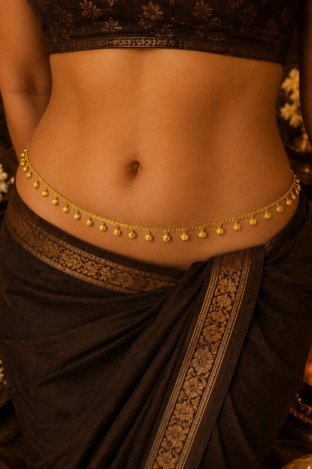 A close-up of a model's waist wearing a delicate gold chain belly chain (kara) with small hanging beads, paired with a dark brown saree with gold borders.