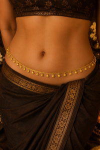 A close-up of a model's waist wearing a delicate gold chain belly chain (kara) with small hanging beads, paired with a dark brown saree with gold borders.