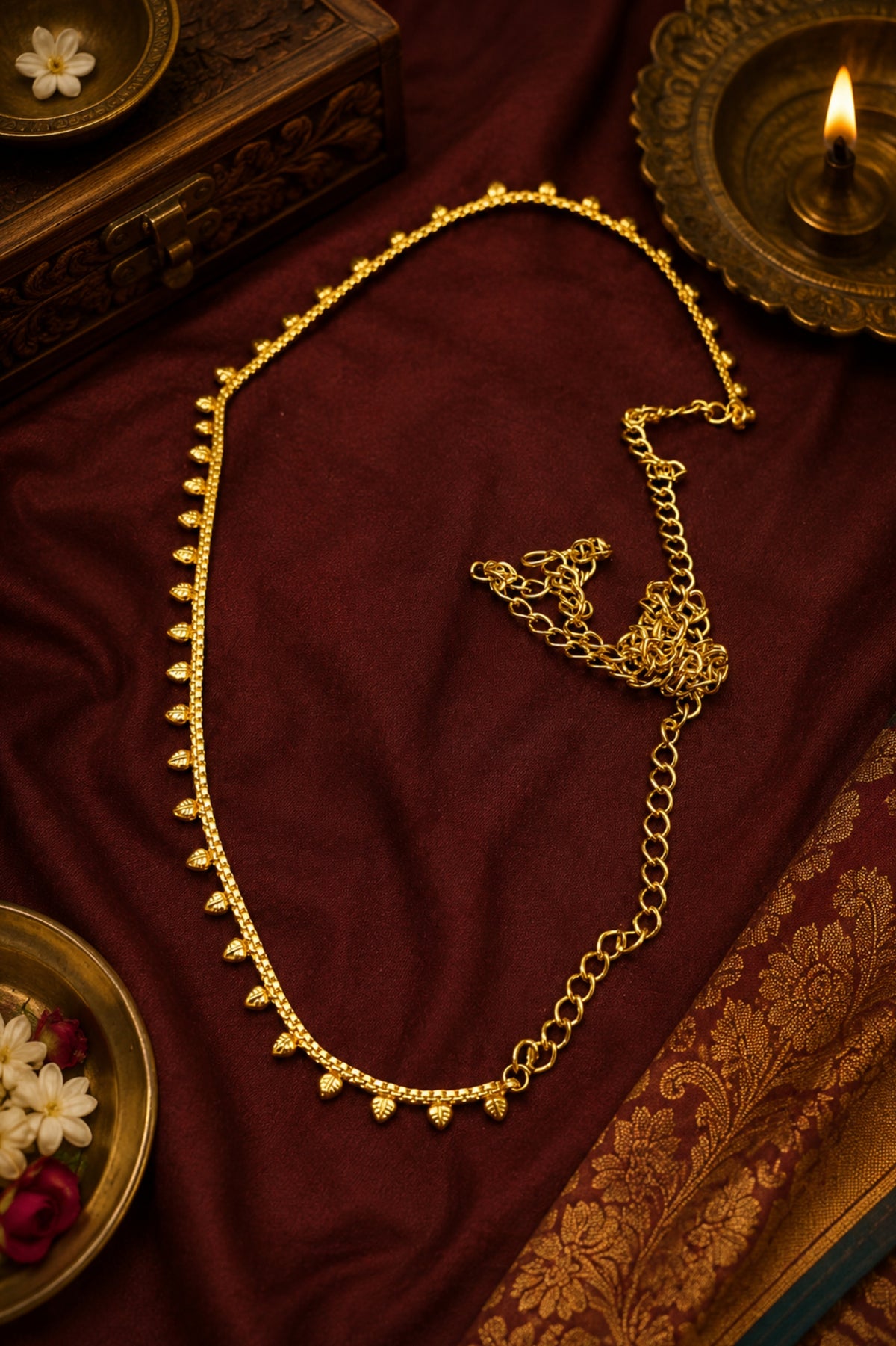 A gold-toned chain necklace with small dangling bead-like accents, arranged on draped deep red fabric with decorative props including a lit oil lamp, carved wooden box, and floral dish.