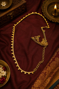A gold-toned chain necklace with small dangling bead-like accents, arranged on draped deep red fabric with decorative props including a lit oil lamp, carved wooden box, and floral dish.