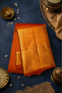 A folded orange and gold zari silk saree displayed on a dark textured blue background with traditional Indian decorative props including brass oil lamps, a diya, and white flowers.